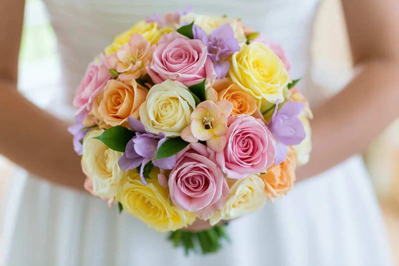 Bride holding a round bouquet of pink, yellow, and orange roses with purple flowers in a white dress