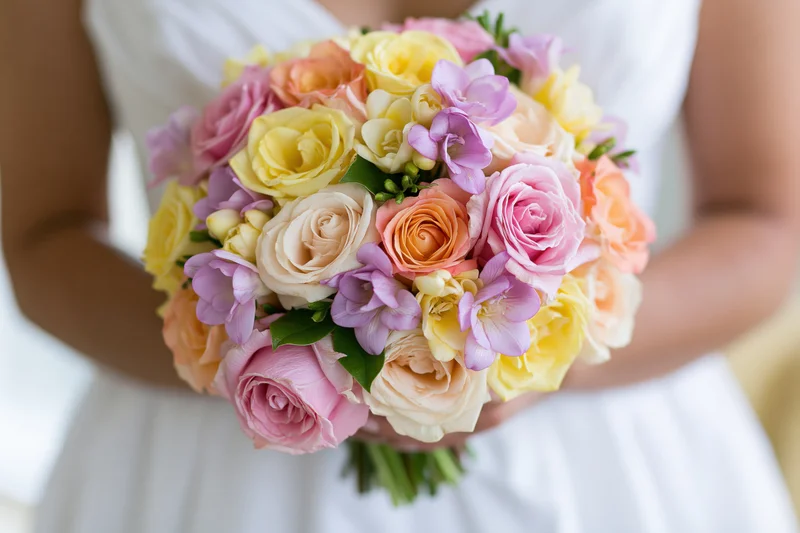 Bride holding a round bouquet of pink, yellow, and peach roses with purple flowers