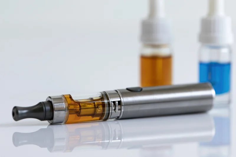 Brushed metal vape pen lying on its side with a clear tank and amber liquid, on a reflective white surface with two e-liquid bottles in the background