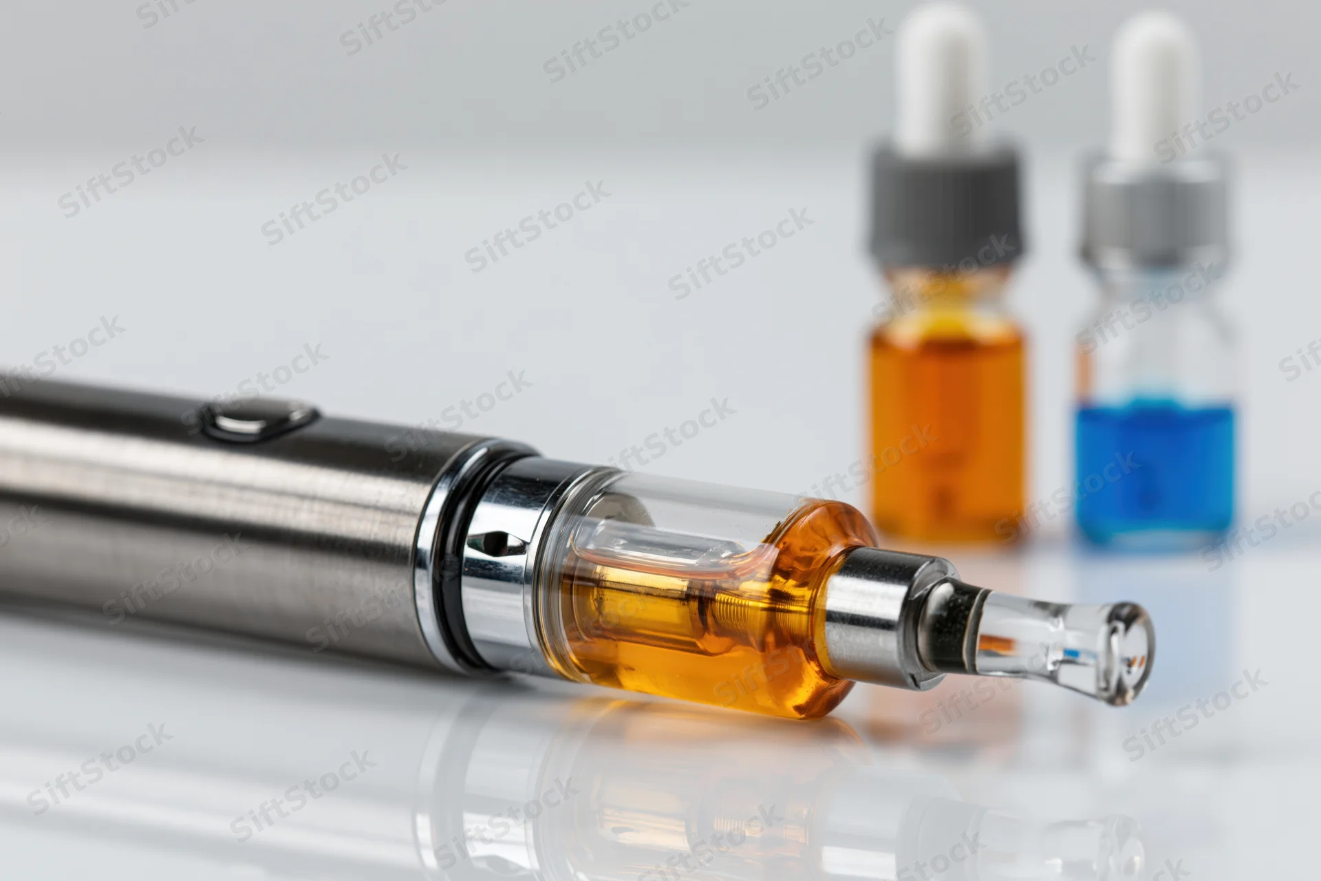 Brushed metal vape pen lying on its side with clear tank and amber liquid, beside two small bottles with droppers on a reflective white surface