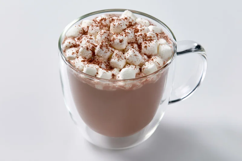 Clear glass double-wall mug filled with hot chocolate topped with marshmallows and cocoa powder on white background