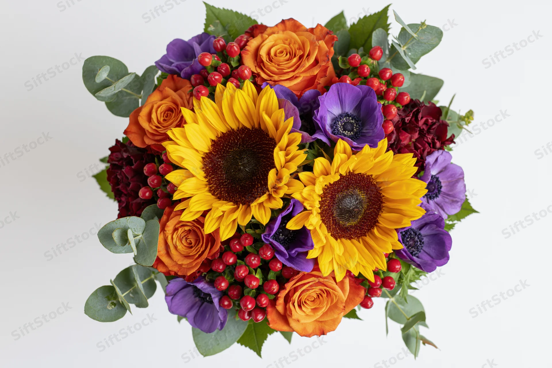 Dense round bouquet of mixed flowers including sunflowers, roses, anemones, eucalyptus, and red berries arranged for display