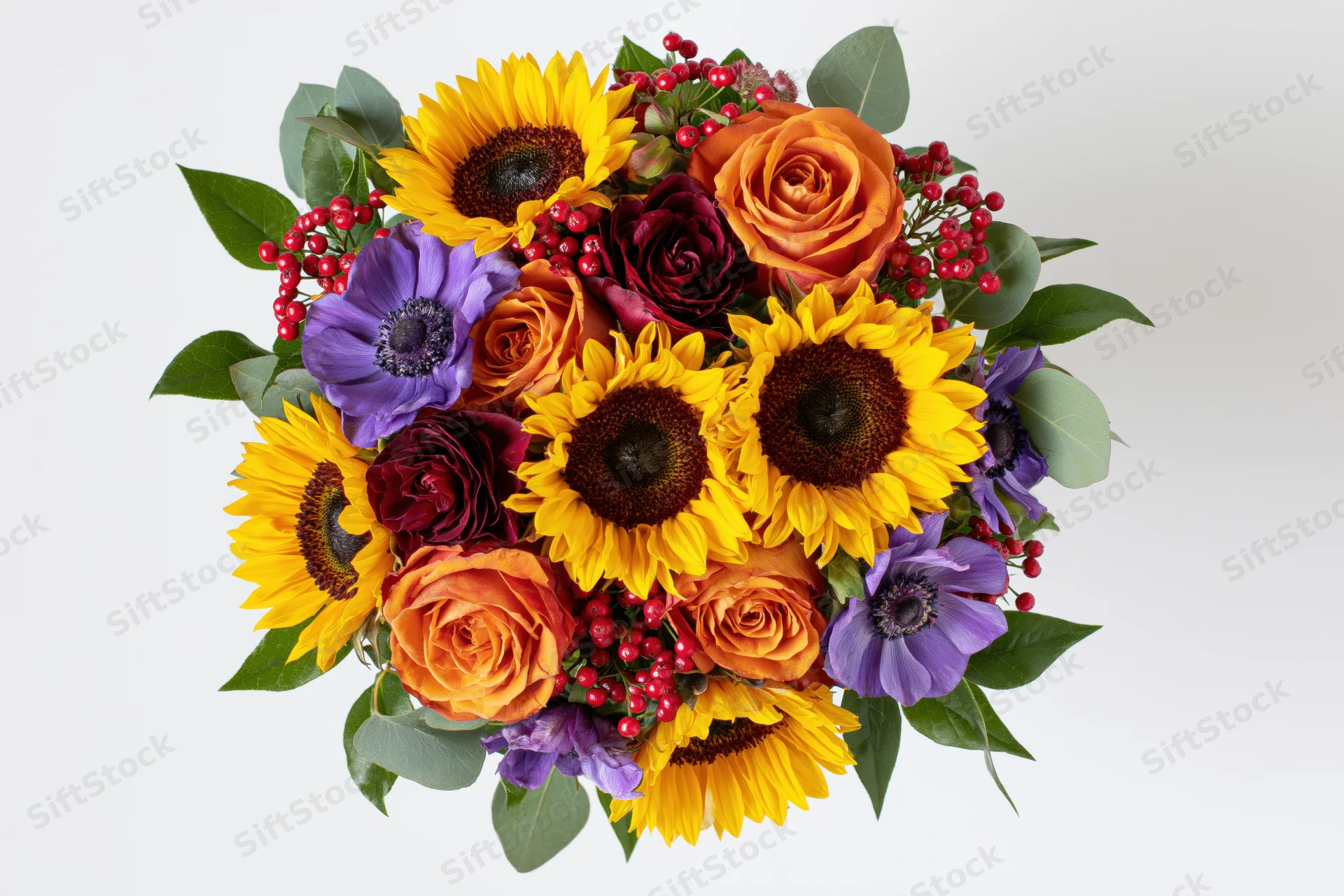 Dense round bouquet of sunflowers, roses, anemones and berries with greenery on white background