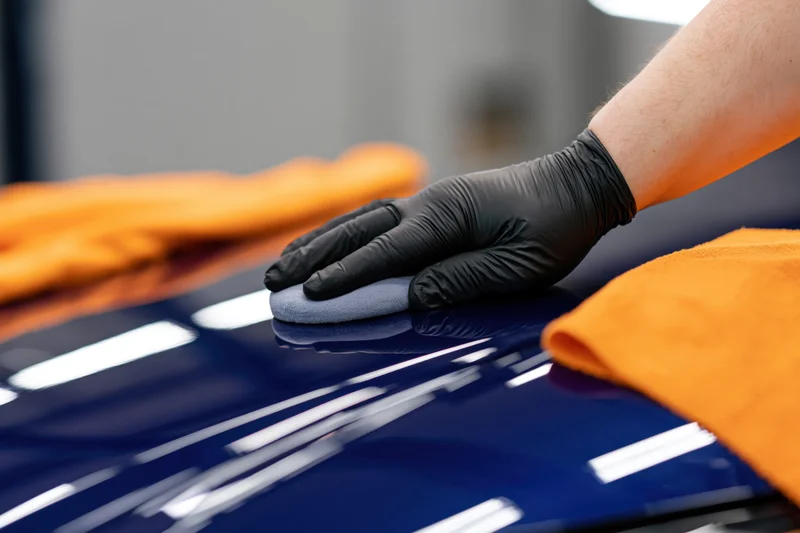 A detailer’s gloved hand applying polish to a glossy blue car surface with a gray applicator pad