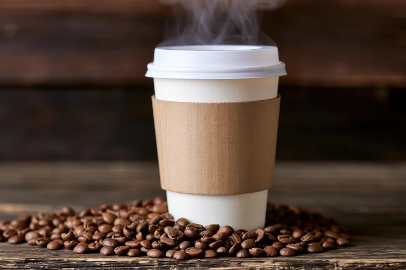 Steaming disposable paper coffee cup with brown sleeve and white lid surrounded by roasted coffee beans on wooden surface
