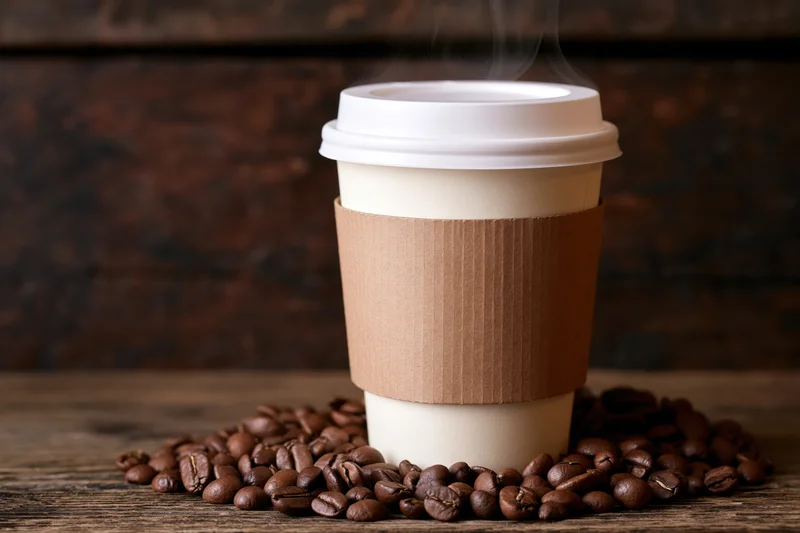 Disposable paper coffee cup with white lid and brown sleeve surrounded by roasted coffee beans on wooden surface