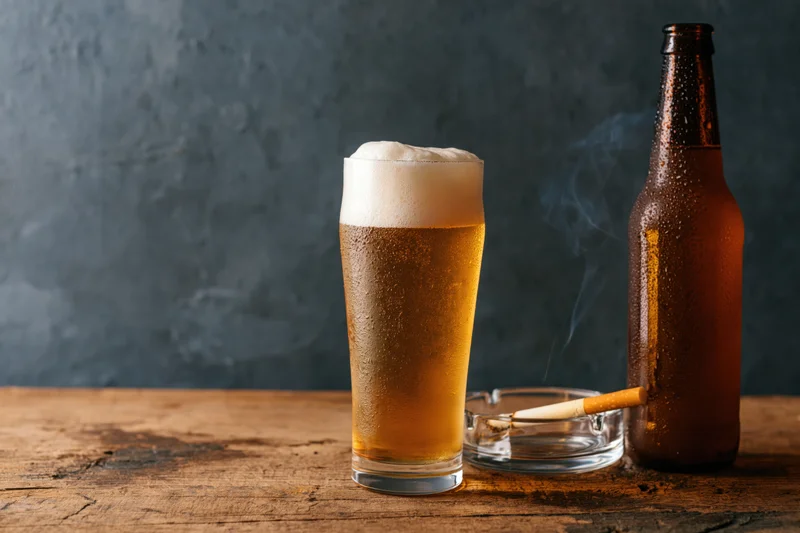 A glass of lager with thick foam and a smoking cigarette in an ashtray beside a bottle on a wooden table