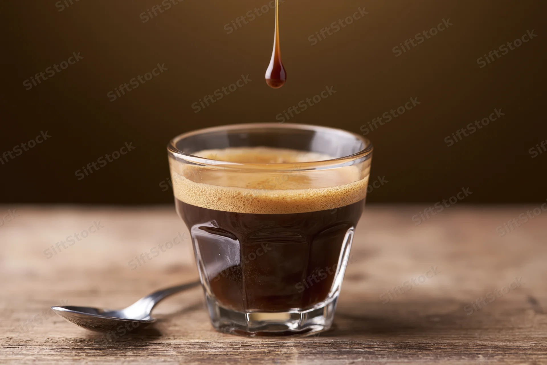 Espresso shot in a glass cup with crema and a drop falling into it on a wooden table beside a spoon