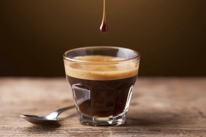 Espresso shot in a glass cup with crema and a drop falling into it on a wooden table beside a spoon