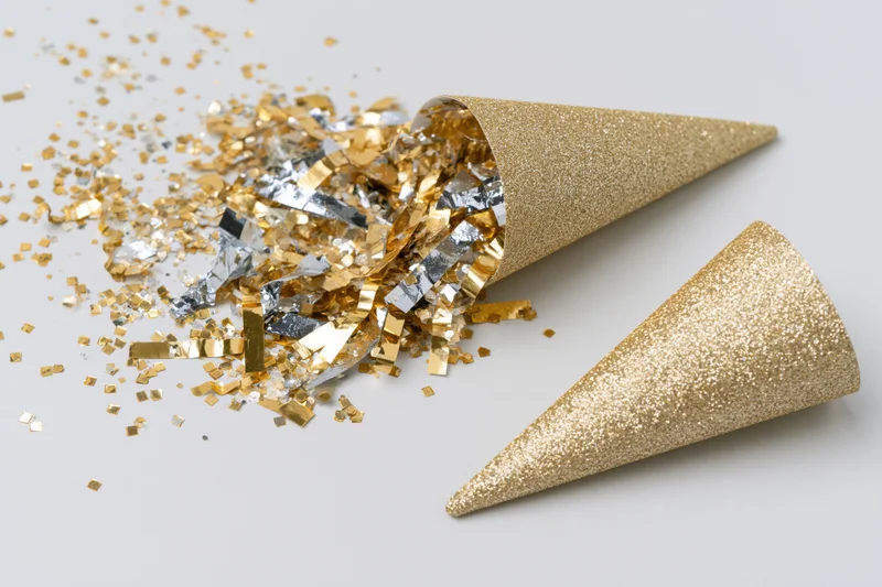 Gold glitter party cone lying on its side with scattered gold and silver confetti on white surface