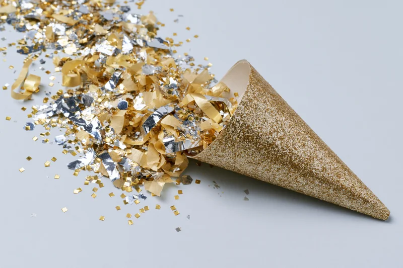 Gold glitter party cone lying on its side with scattered gold and silver confetti on a plain surface
