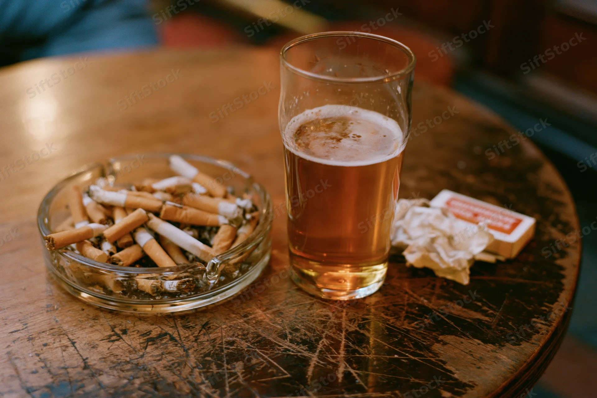 Half-full pint glass of lager beside an overfilled glass ashtray with cigarette butts on a scratched wooden table