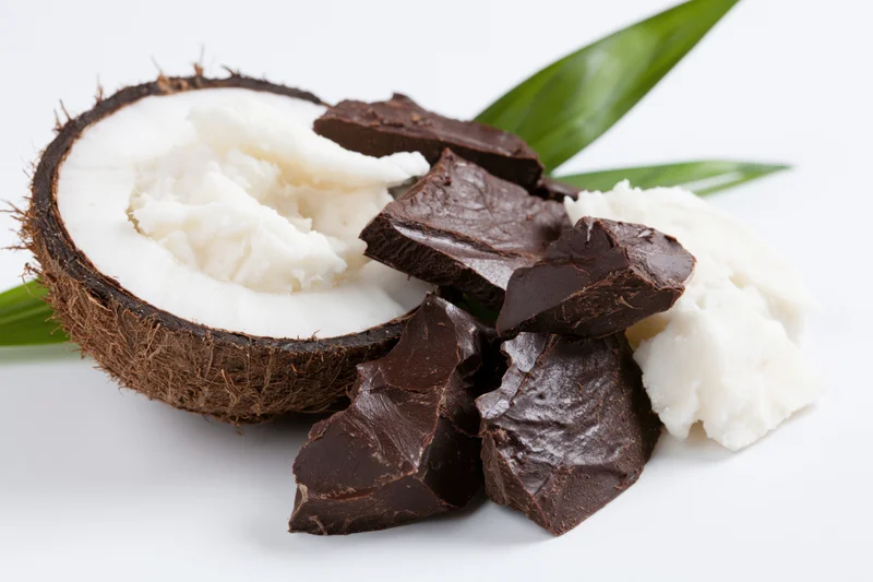 Halved coconut with white flesh and broken dark chocolate pieces on white background with green leaf