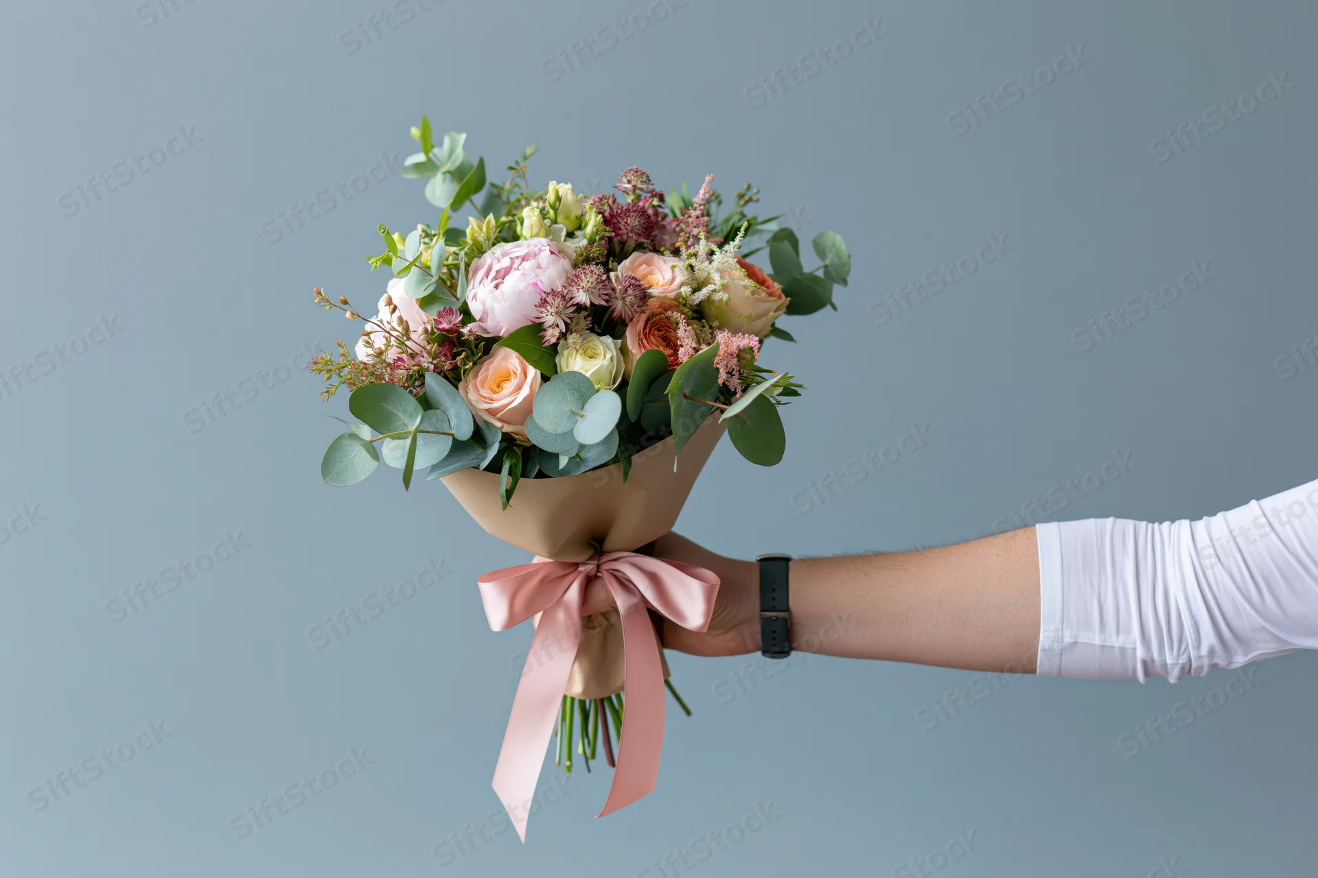 Hand extending a wrapped bouquet of roses and peonies with greenery and pink ribbon against a plain background