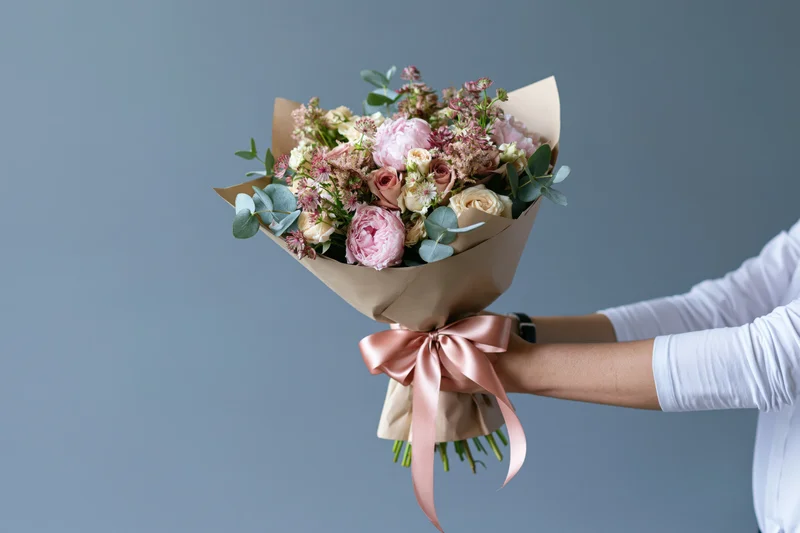Woman extending a wrapped bouquet of pink peonies, roses and eucalyptus against a plain background