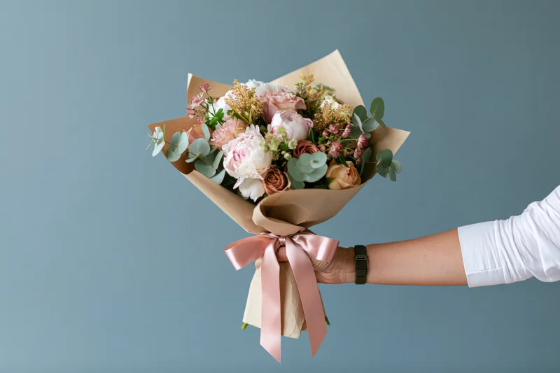 Woman's hand holding a wrapped bouquet of peonies and roses with pink ribbon against a plain background