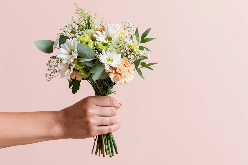 Hand holding a mixed flower bouquet with white daisies, peach carnations, and greenery against a pink background