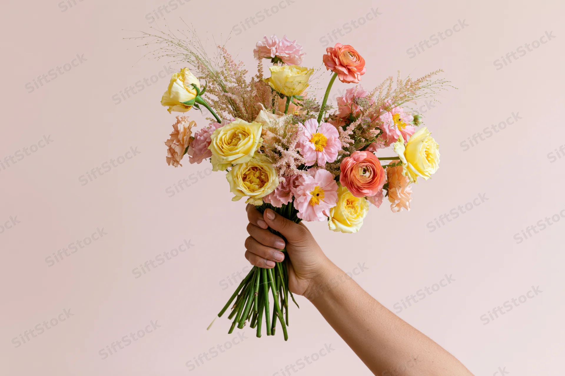 Hand holding a loose mixed bouquet of yellow pink and peach flowers against a soft pink background