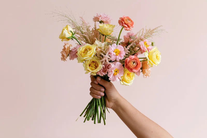 Hand holding a loose mixed bouquet of yellow pink and peach flowers against a soft pink background