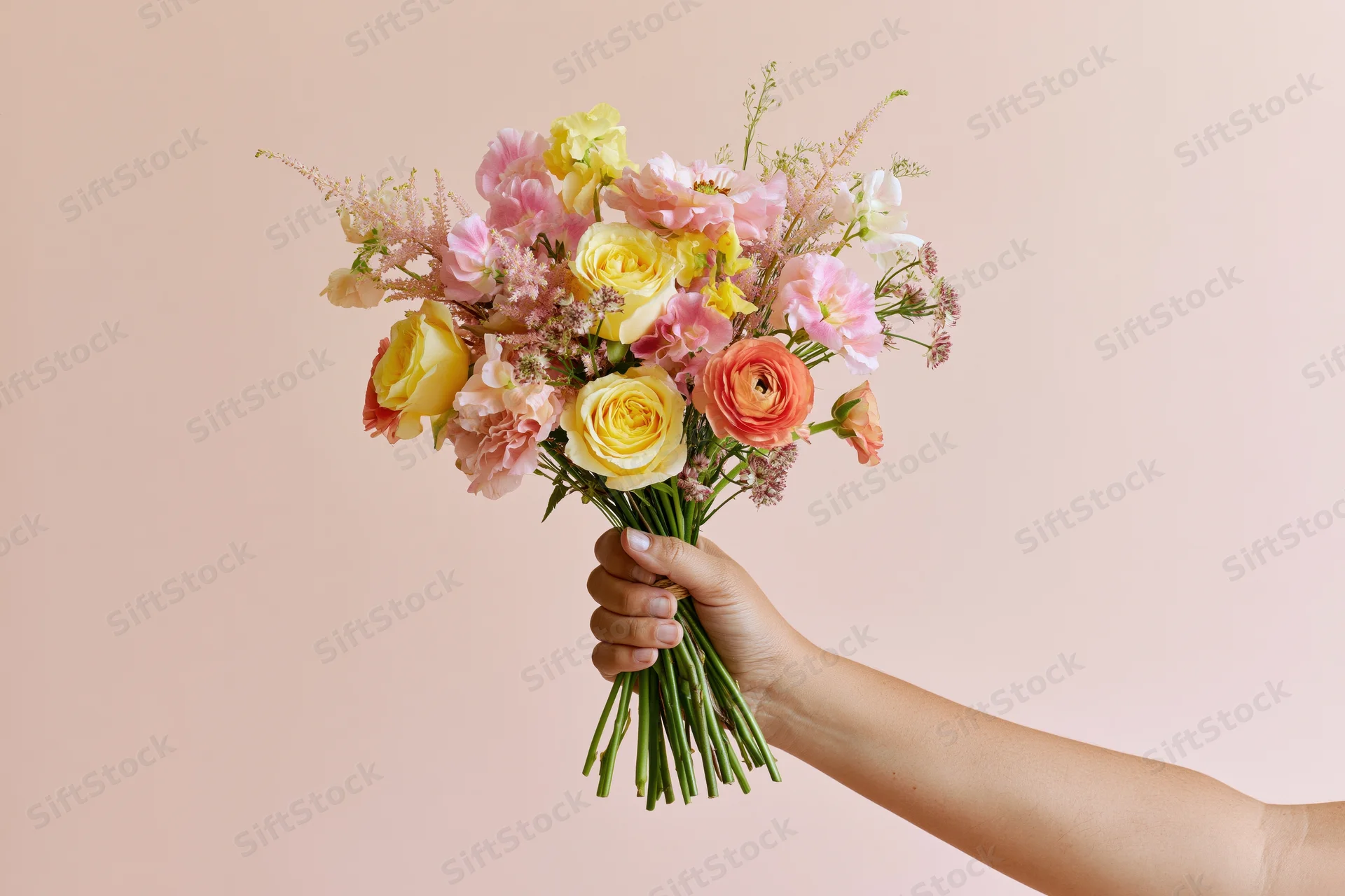 Woman holding a loose mixed flower bouquet with yellow roses and pink ranunculus against a soft pink background