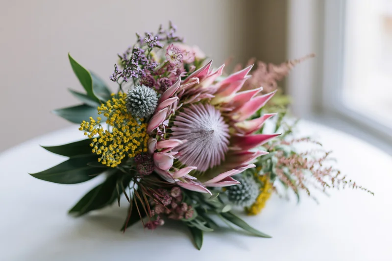 King protea at center of mixed wildflower bouquet on white table near window