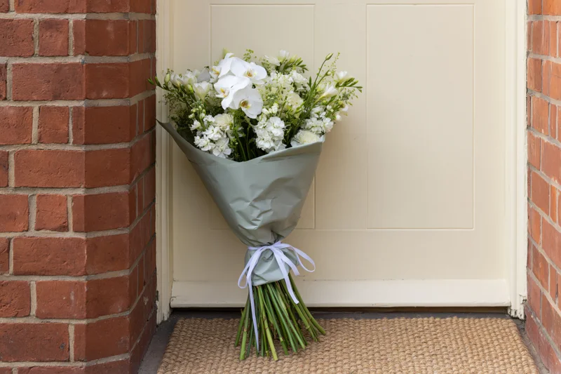 White flower bouquet wrapped in paper with ribbon resting on doormat at front door entrance