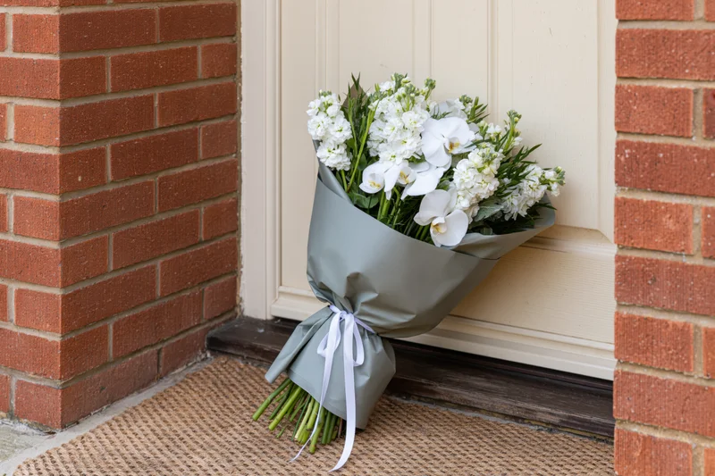 Wrapped white flower bouquet resting on doormat beside brick house entrance