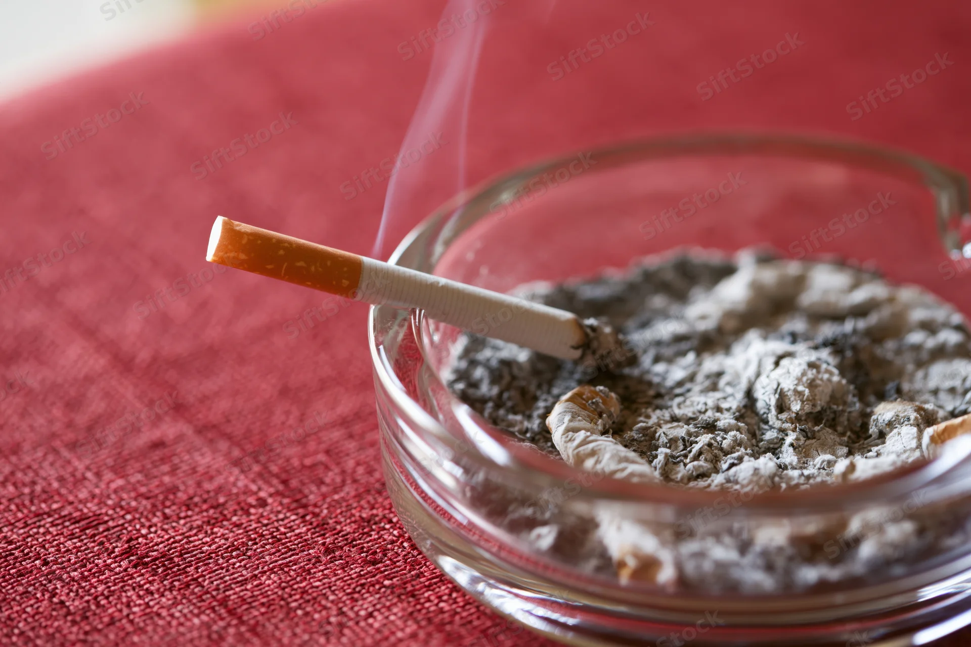 Lit cigarette resting in glass ashtray filled with ash on red tablecloth