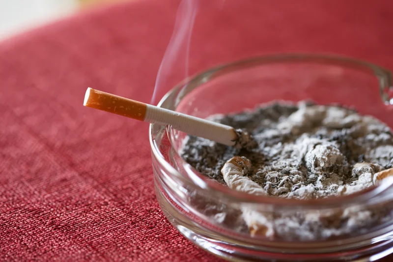 Lit cigarette resting in glass ashtray filled with ash on red tablecloth