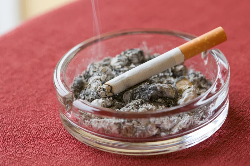 Lit cigarette resting in glass ashtray filled with ash on red tablecloth