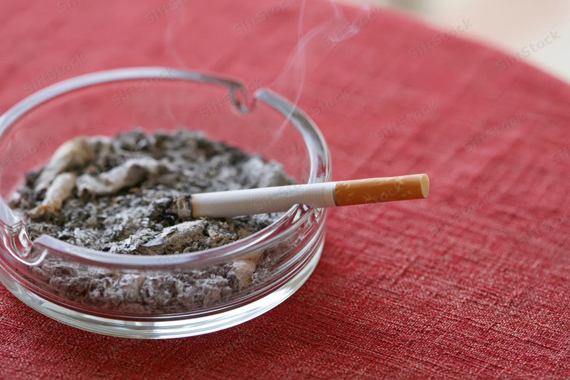 Lit cigarette resting in glass ashtray filled with ash on red tablecloth