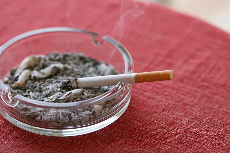 Lit cigarette resting in glass ashtray filled with ash on red tablecloth
