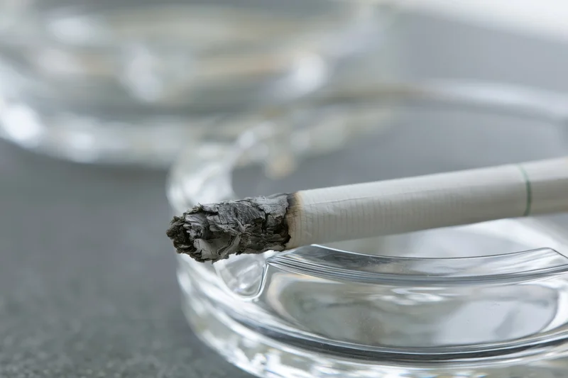 Lit cigarette with long gray ash resting on glass ashtray