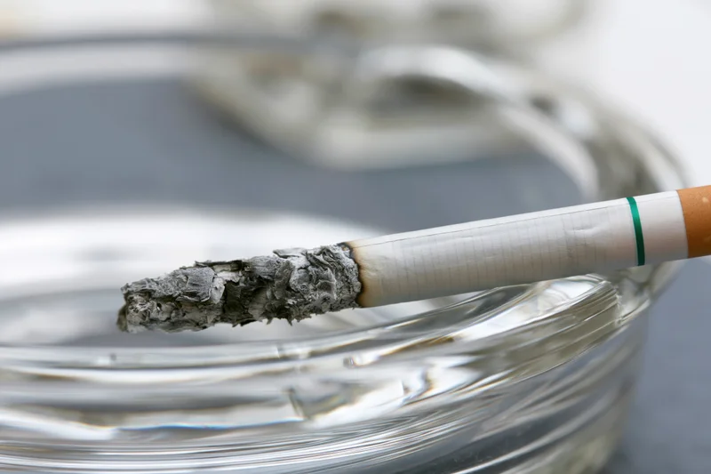 Lit cigarette with long gray ash resting across grooved glass ashtray