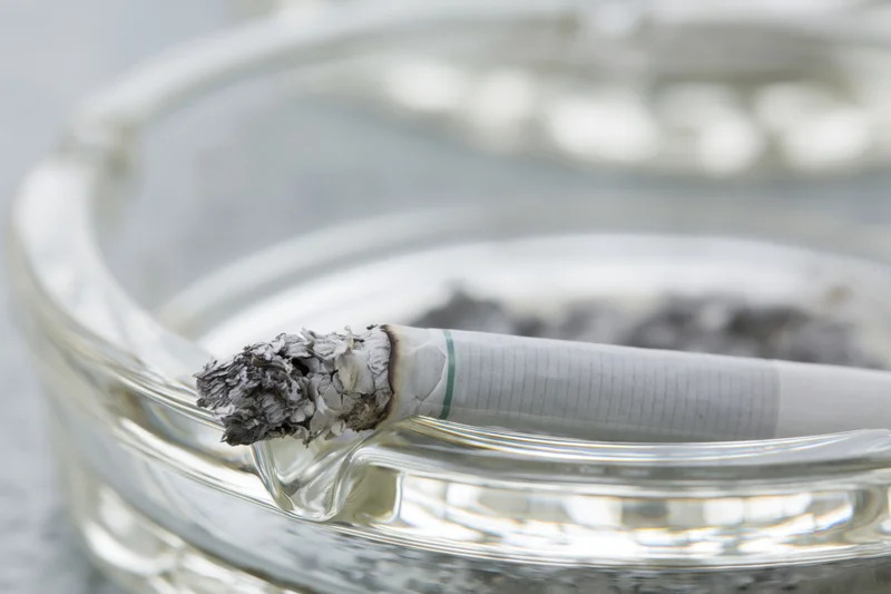 Lit cigarette with long gray ash resting horizontally across glass ashtray groove