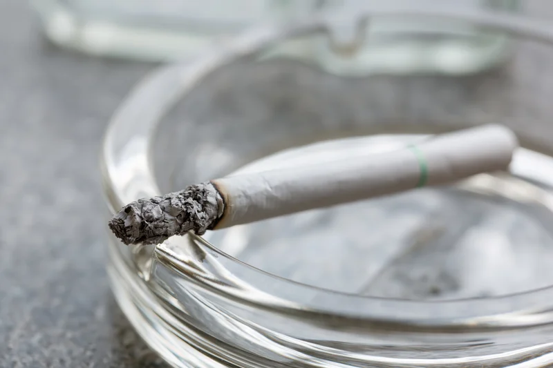 Lit cigarette with long gray ash resting across glass ashtray rim