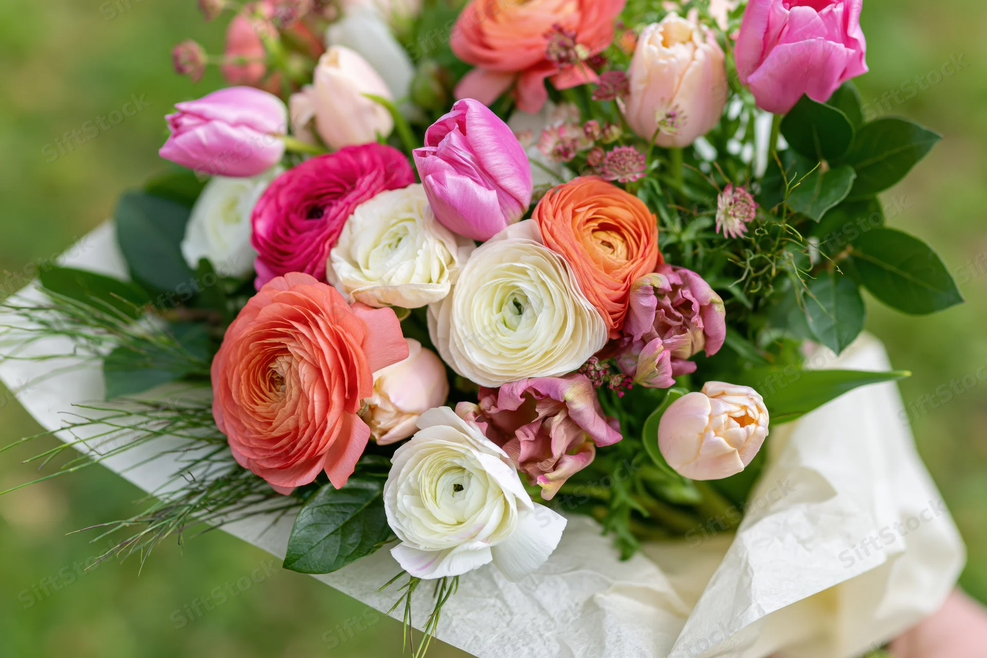 A handheld mixed bouquet of ranunculus and tulips with greenery wrapped in paper