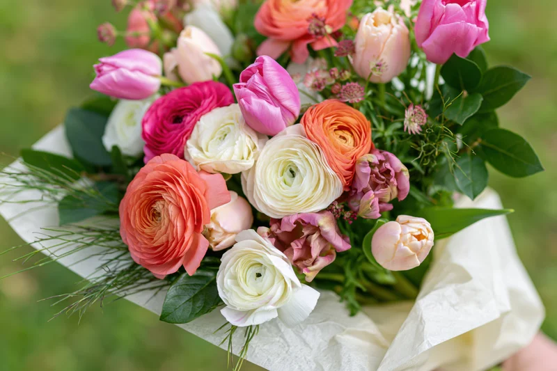 A handheld mixed bouquet of ranunculus and tulips with greenery wrapped in paper