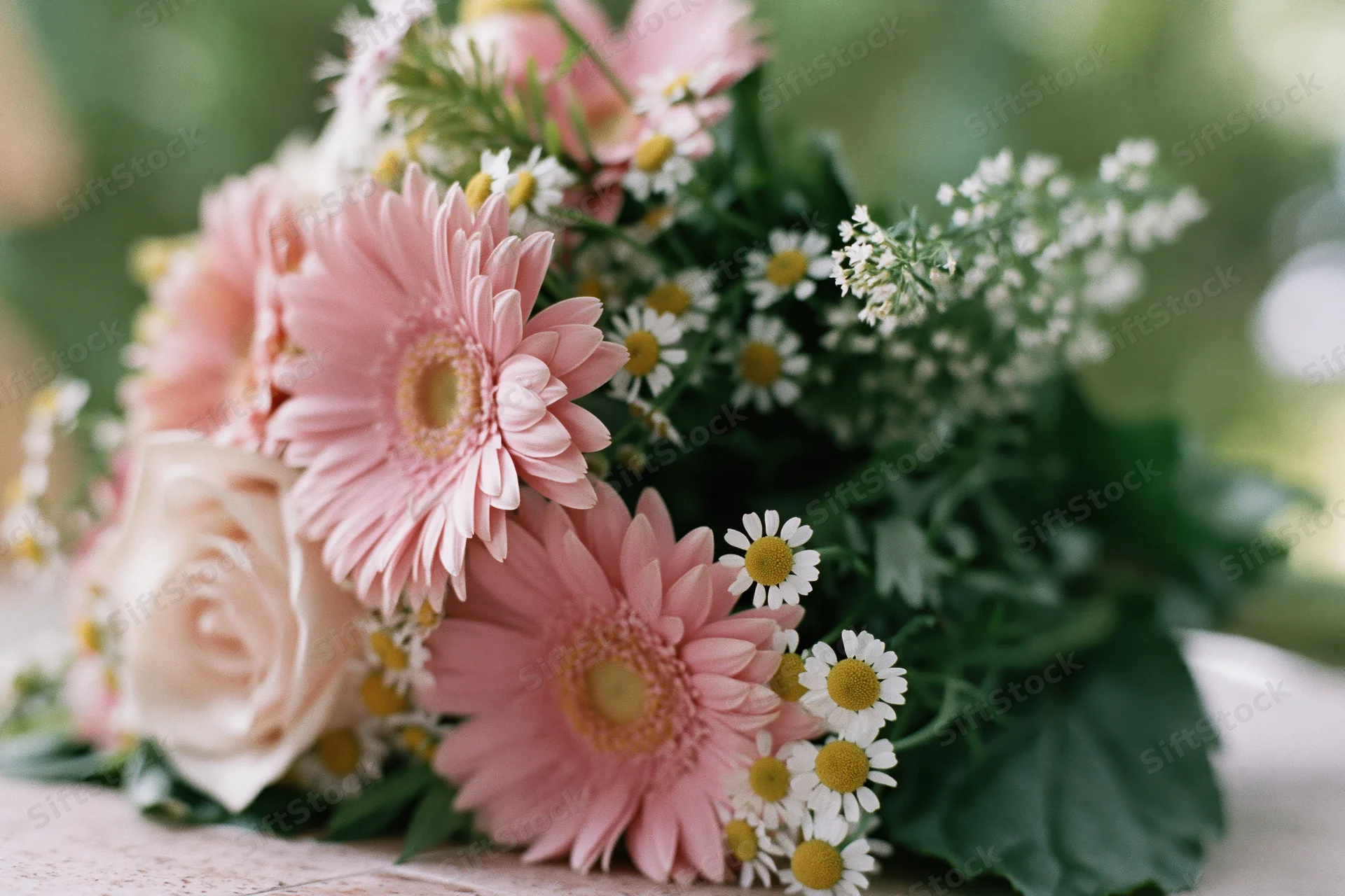 Mixed flower bouquet with pink georginas, roses, and daisies resting on a surface with green foliage and soft background