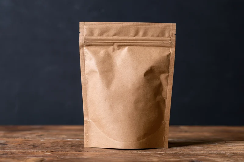 Plain kraft paper zip-lock pouch standing upright on a worn wooden table against a dark background