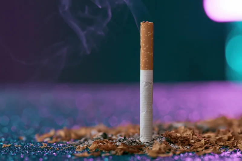 Single cigarette burning upright on glittery surface with smoke rising and colorful bokeh background