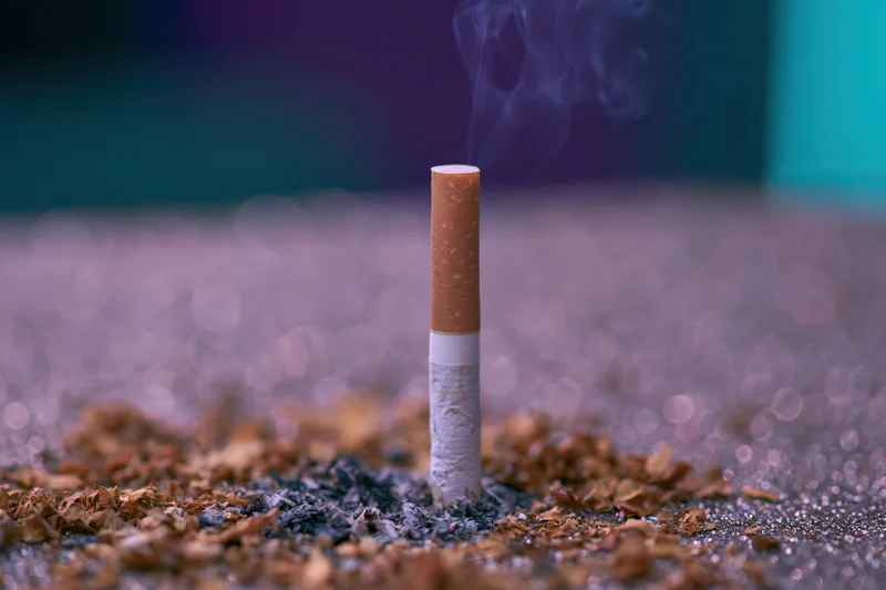 Single cigarette burning upright on a glittery surface with smoke rising and ash scattered around