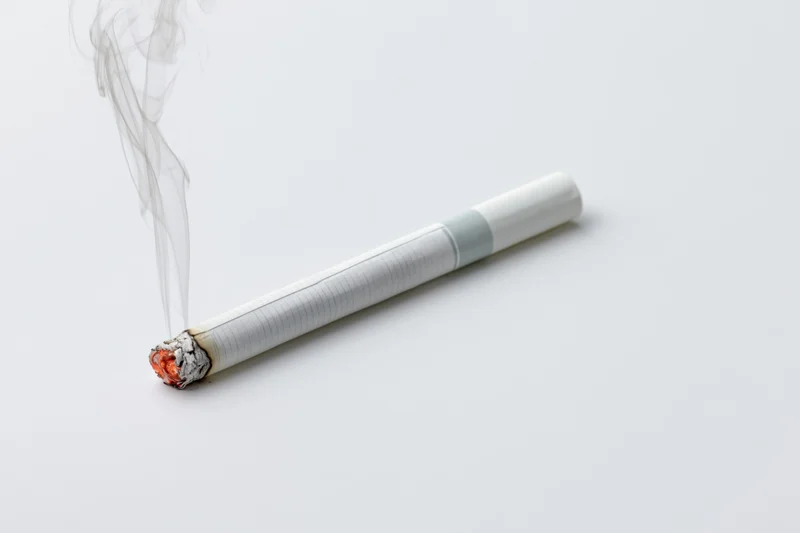 Lit cigarette burning horizontally with glowing ember and rising smoke on white surface
