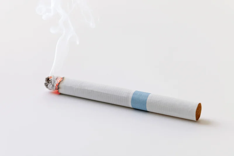 Lit cigarette resting horizontally with thin white smoke rising from burning end on white background