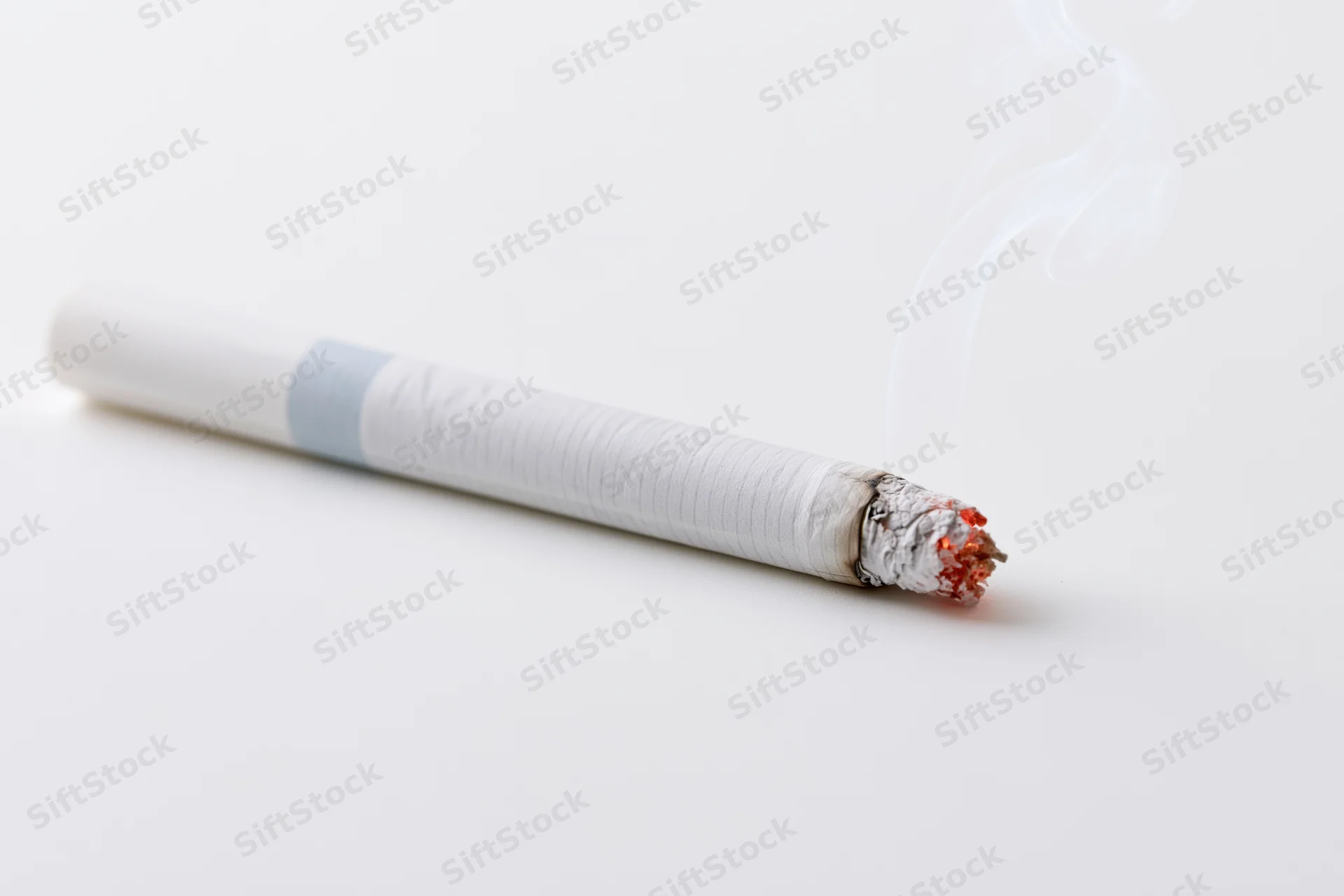 Lit cigarette resting horizontally with thin white smoke rising from burning end on white surface