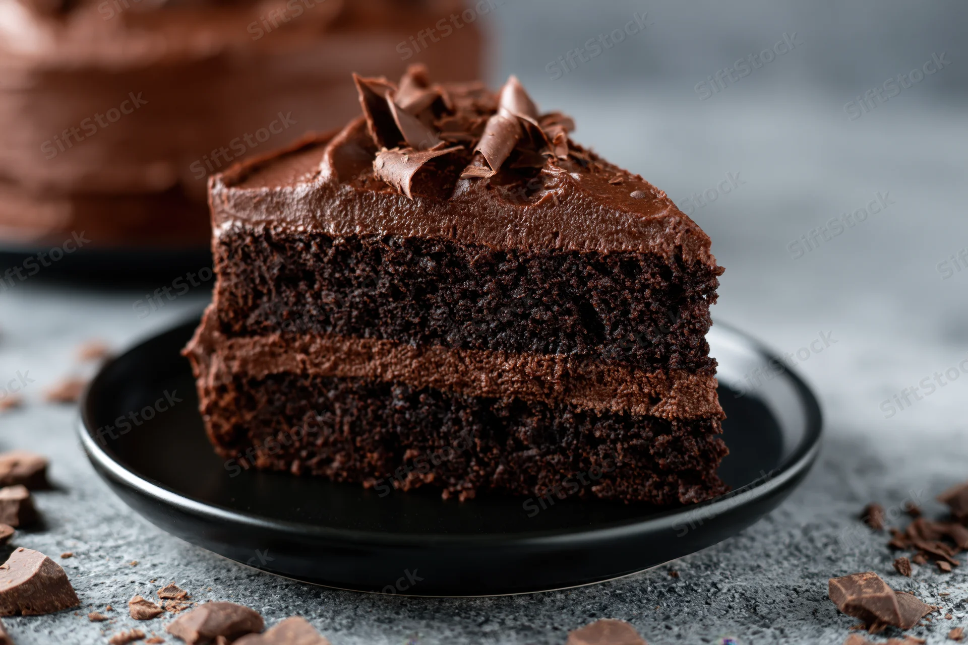 Slice of triple-layer dark chocolate cake with thick ganache frosting on black plate surrounded by chocolate chunks