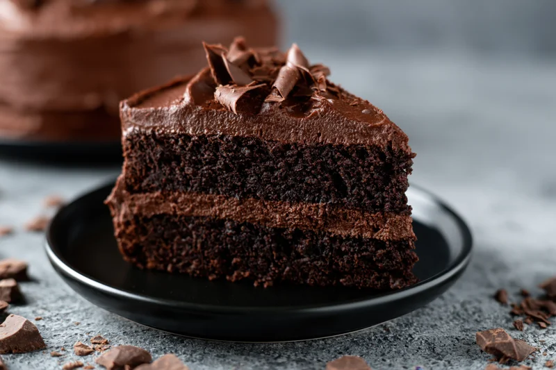 Slice of triple-layer dark chocolate cake with thick ganache frosting on black plate surrounded by chocolate chunks