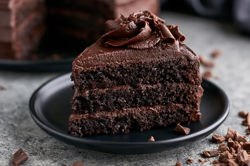 Slice of triple-layer dark chocolate cake with thick ganache frosting on black plate