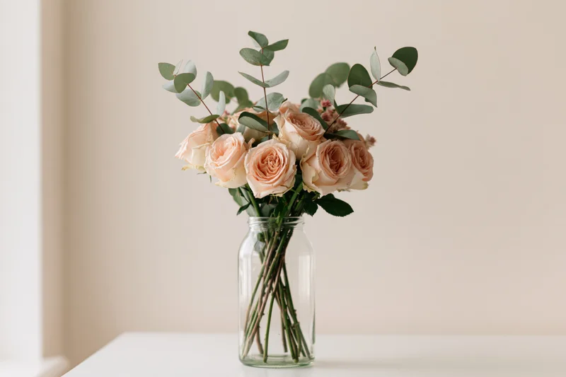 Peach roses and eucalyptus arranged in a glass vase on a white table