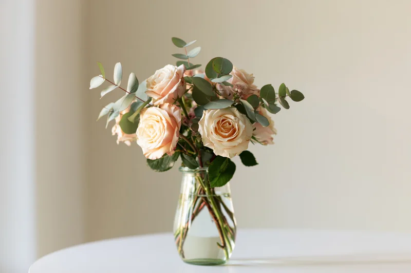 Peach roses and eucalyptus arranged in a glass vase on a white table